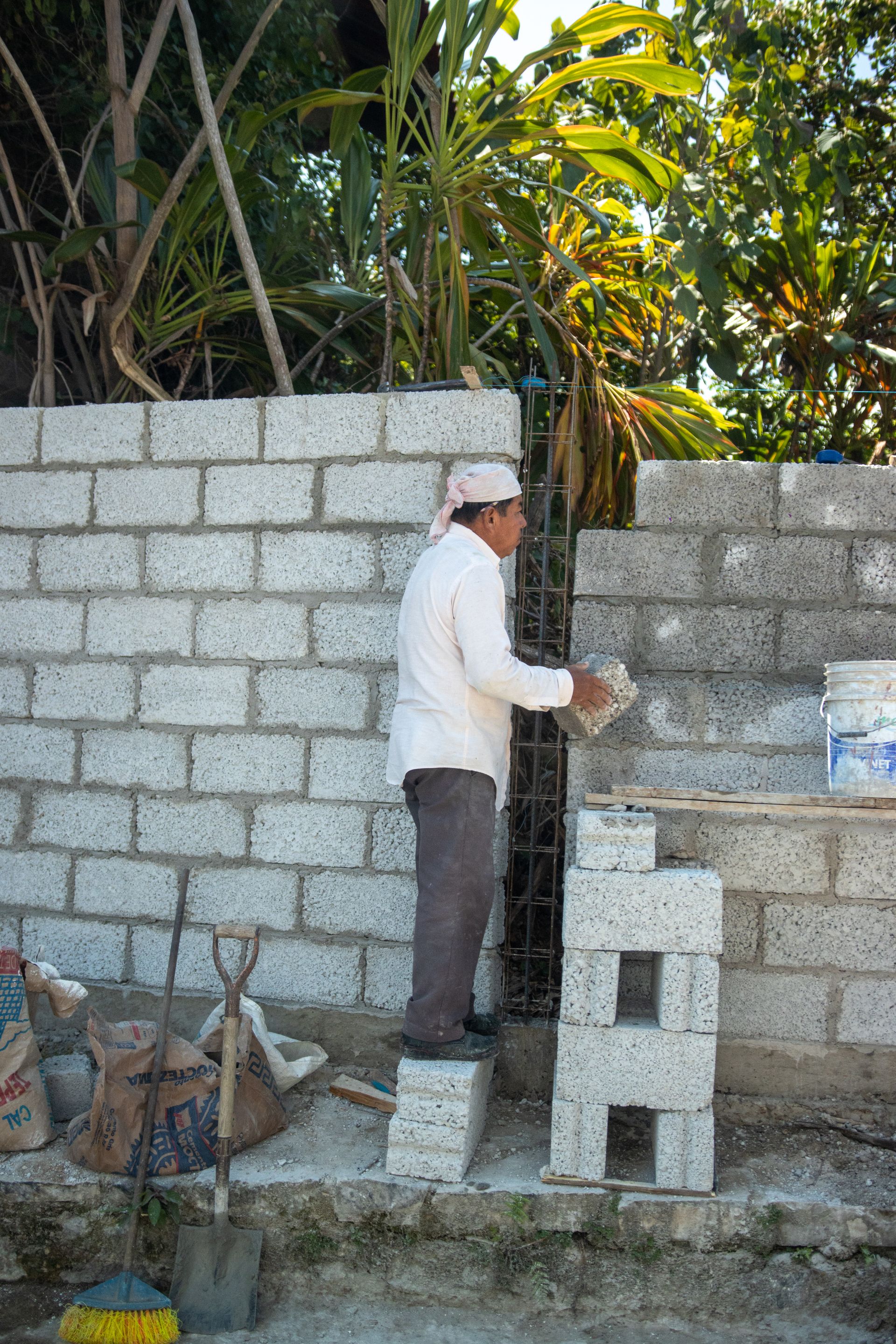A construction worker building a concrete block wall surrounded by tropical vegetation