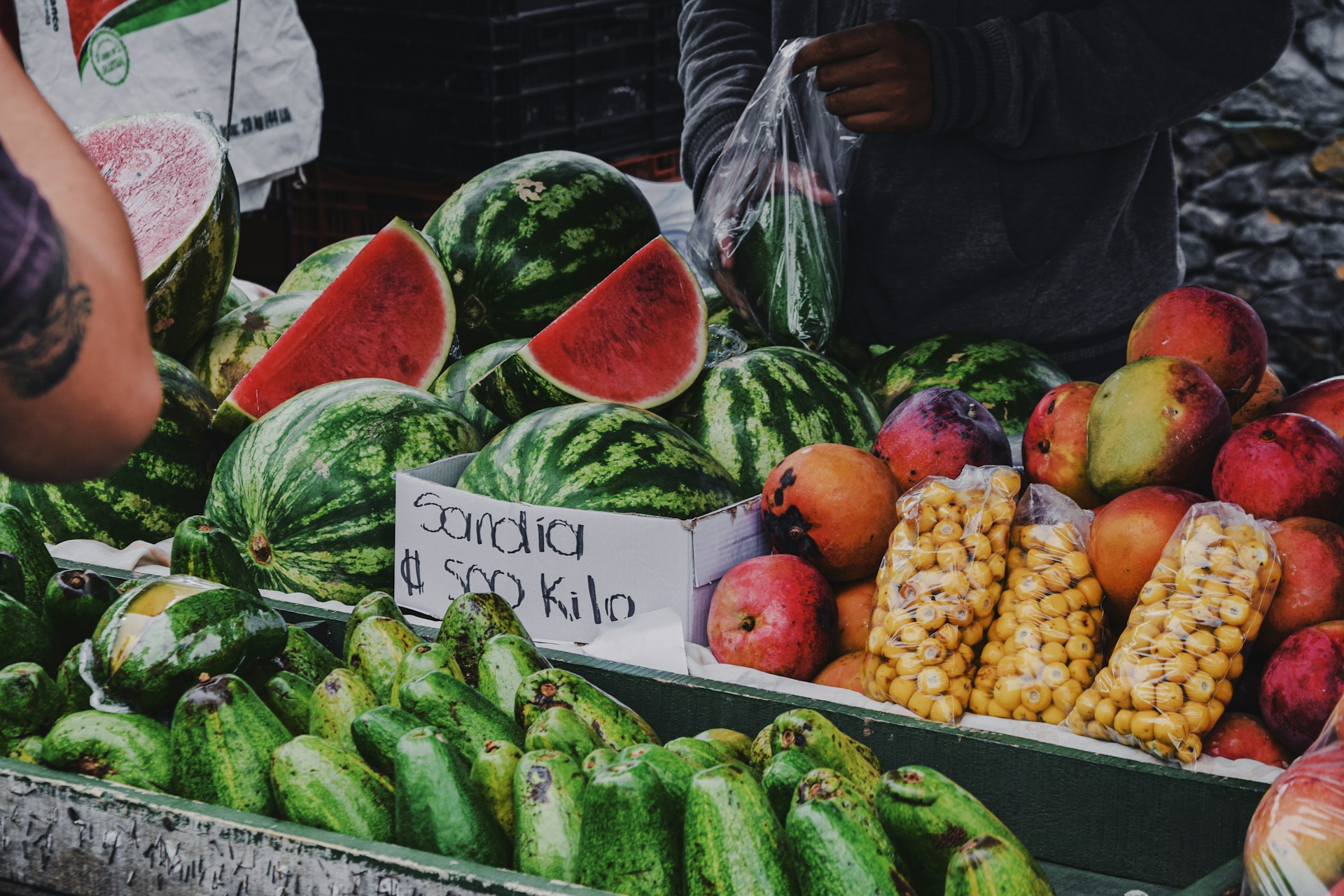 A Costa Rican farmers' market stall with watermelons, mangoes and nances, and a handwritten price sign in colones