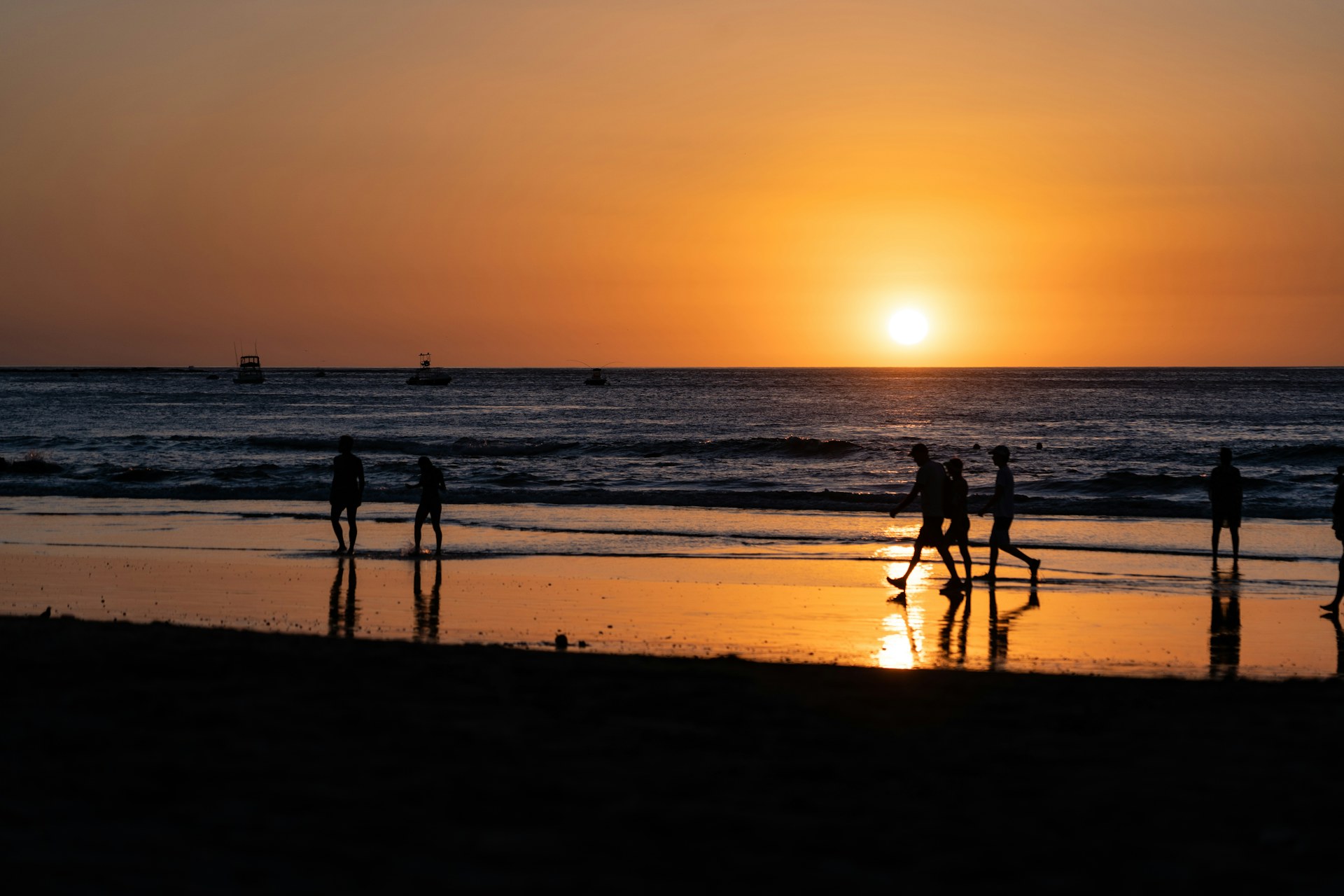 People walking along a Pacific beach at sunset, with boats anchored offshore